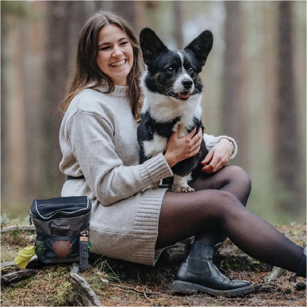 Frauchen sitzt mit Ihrem Hund auf dem Schoß im Wald und hat die Rudelkönig Gassitasche mit sich, um den Hund perfekt zu trainieren.