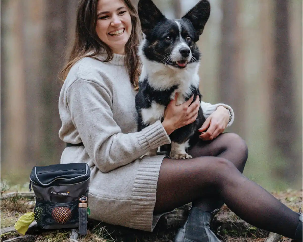 Frauchen sitzt mit Ihrem Hund auf dem Schoß im Wald und hat die Rudelkönig Gassitasche mit sich, um den Hund perfekt zu trainieren.