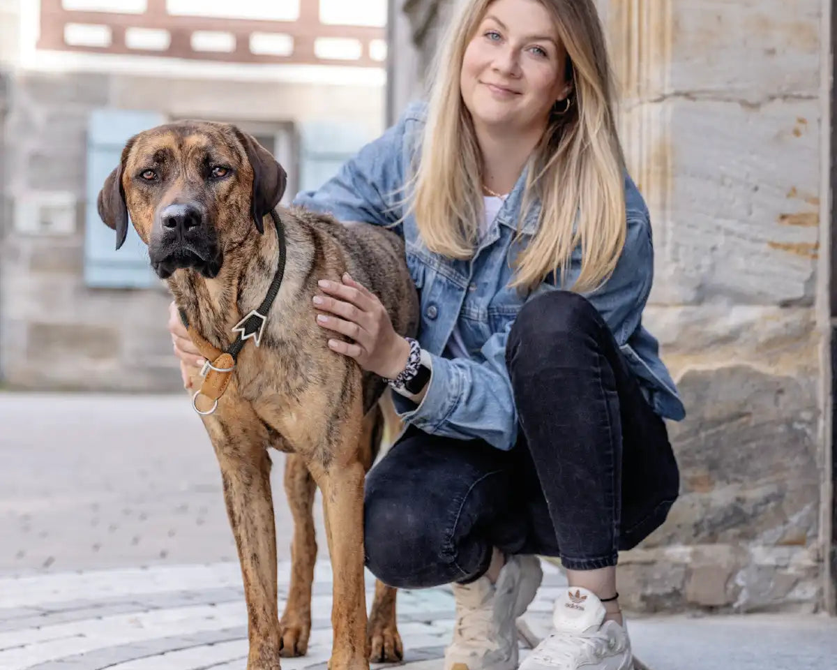 Frauchen steht kniet mit ihrem Hund und dem Rudelkönig Zugstophalsband Emma in der Stadt und beide schauen in die Kamera.
