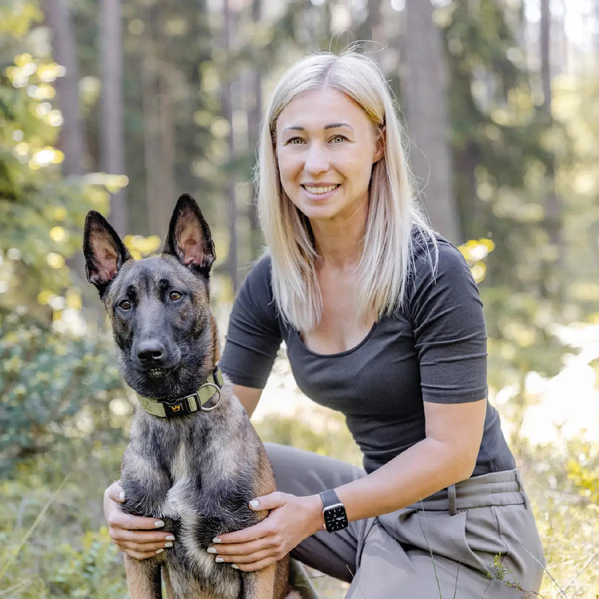 Frau mit blondem Haar kniet im Wald neben ihrem Hund und hält ihn liebevoll fest. Der Hund trägt ein olivgrünes Halsband Bella von Rudelkönig und blickt ruhig in die Kamera - harmonische Szene zwischen Mensch und Hund in der Natur.