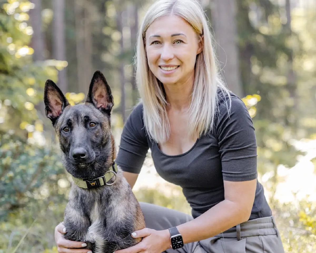 Frau mit blondem Haar kniet im Wald neben ihrem Hund und hält ihn liebevoll fest. Der Hund trägt ein olivgrünes Halsband Bella von Rudelkönig und blickt ruhig in die Kamera - harmonische Szene zwischen Mensch und Hund in der Natur.
