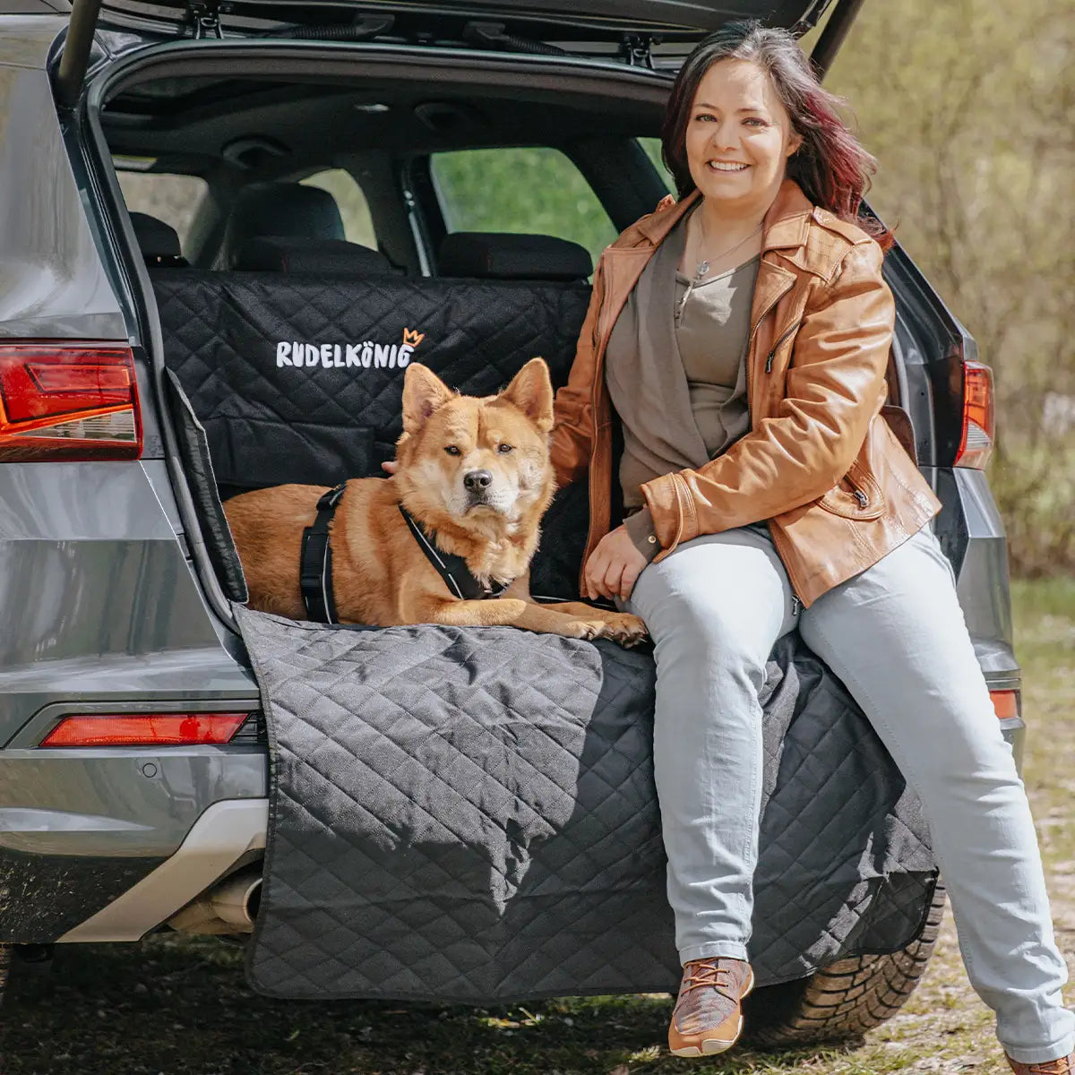 Frau sitzt mit ihrem Hund auf der geöffneten Kofferraumklappe, geschützt durch einen Kofferraumschutz – ideal für Reisen mit Hund.