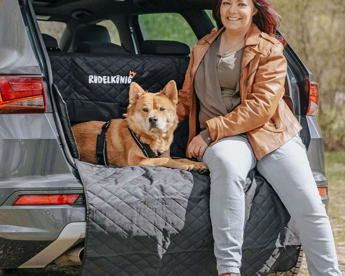 Frau sitzt mit ihrem Hund auf der geöffneten Kofferraumklappe, geschützt durch einen Kofferraumschutz – ideal für Reisen mit Hund.