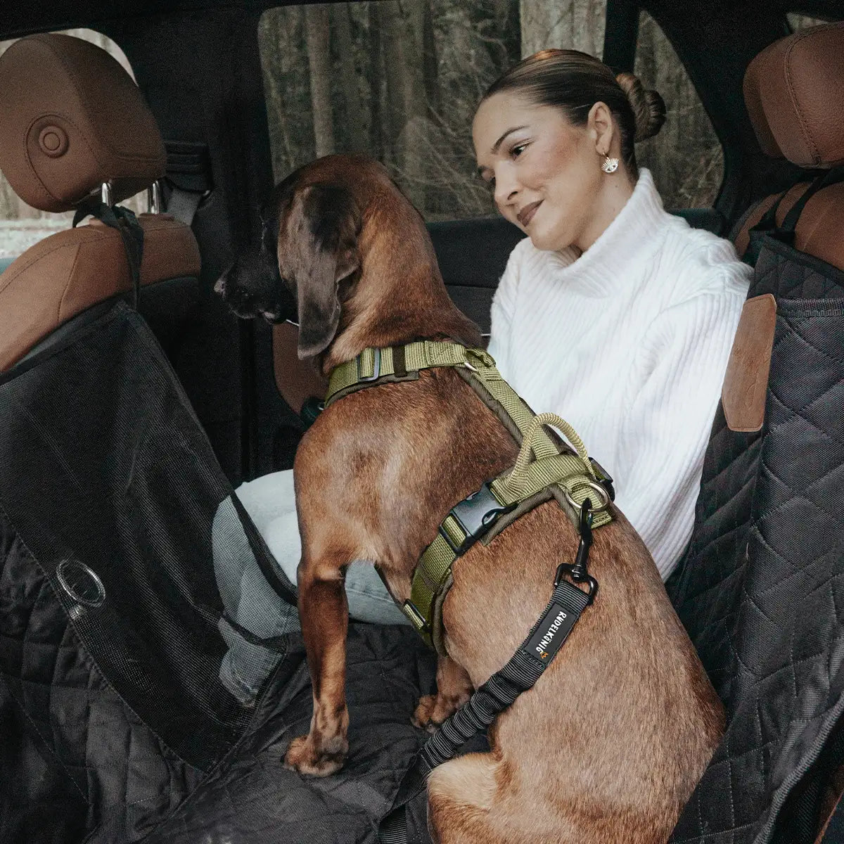 Hund sitzt mit Frauchen auf der Rückbank im Auto, angeleint am Geschirr auf einem Rücksitzschutz – sicher und bequem unterwegs.