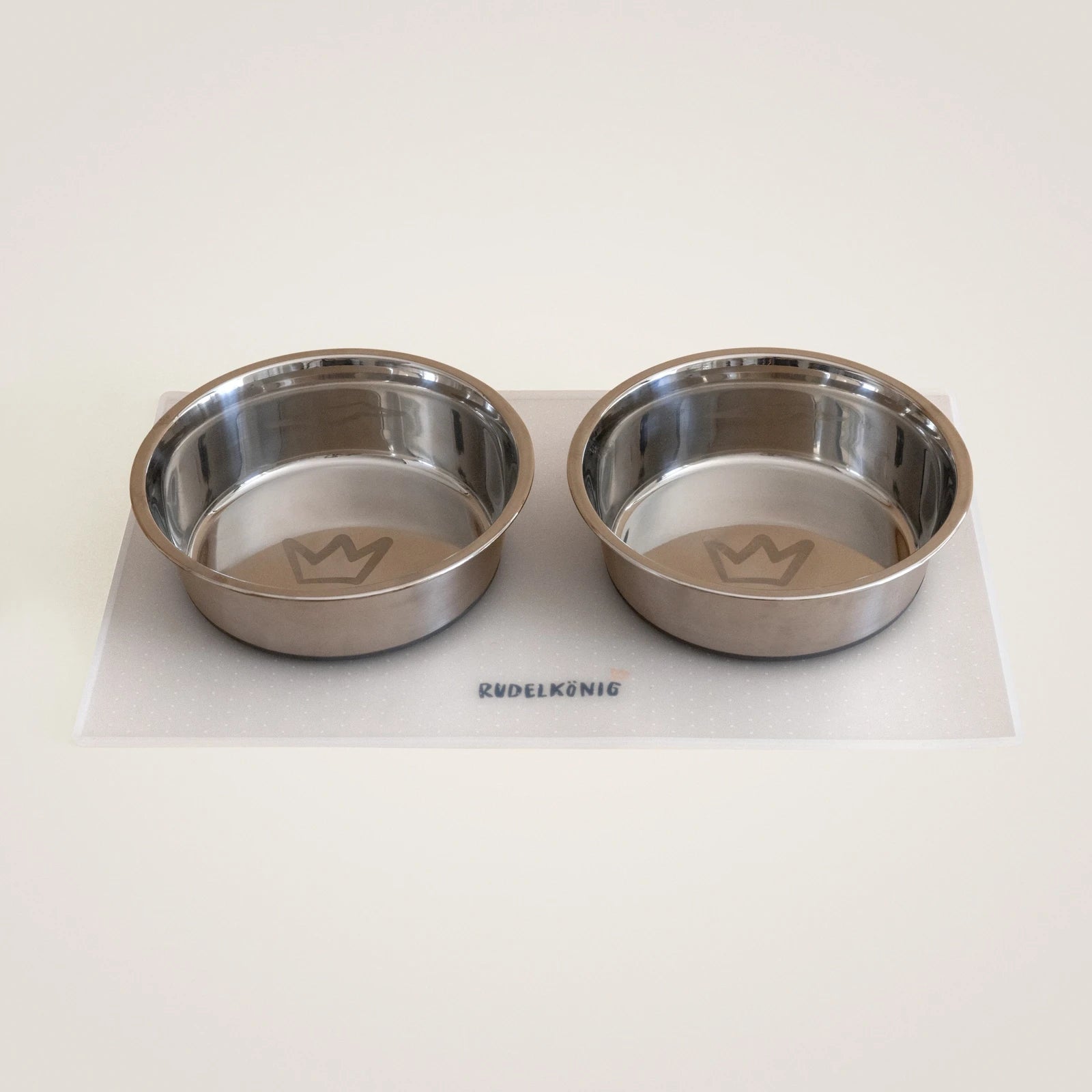 Das Hundenapf Set aus Edelstahl von Rudelkönig mit zwei Hundenäpfen und einer rutschfesten Futtermatte in grau oder transparent.